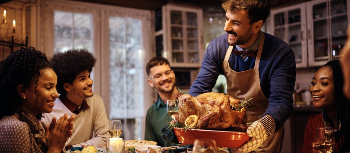 Happy man serving roasted turkey during Thanksgiving dinner with friends at dining table.