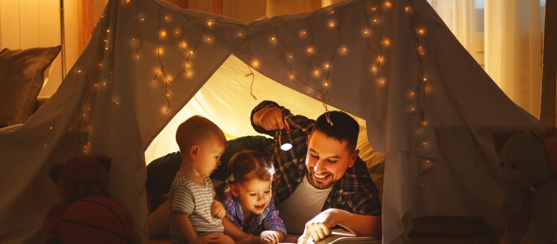 happy family father and children reading a book with a flashlight in a tent at home